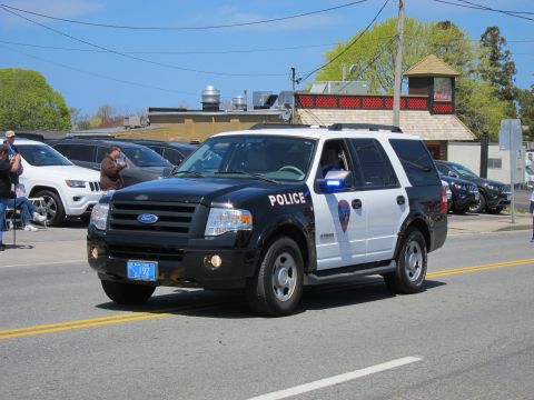 1991 Ford LTD Crown Victoria - black and white Ford Expedition SUV lettered "POLICE" drives down a street with spectators seated along the curb.
