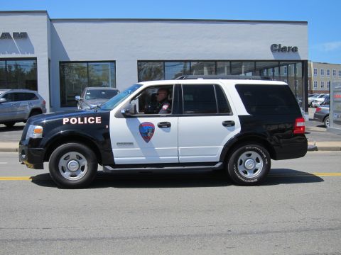 1991 Ford LTD Crown Victoria - black and white Ford Expedition police SUV with a badge decal drives past a dealership storefront.