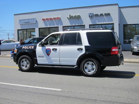 1991 Ford LTD Crown Victoria - black and white Ford Expedition police SUV with badge decal drives past a Jeep and Ram dealership with onlookers.