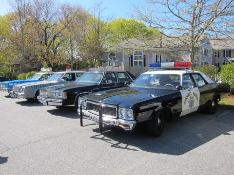 1991 Ford LTD Crown Victoria - lineup of restored vintage police cars, led by a black and white California Highway Patrol sedan, parked on a grassy yard by a house.