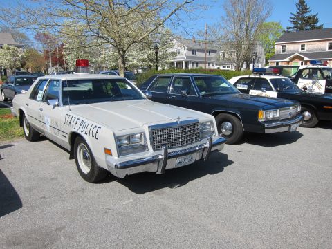 1991 Ford LTD Crown Victoria - white vintage "State Police" sedan parked in a row of classic police cruisers near houses and flowering trees.