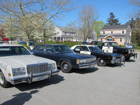 1991 Ford LTD Crown Victoria - row of vintage sedans, including a white police car and black and white Highway Patrol cruisers, parked on pavement near homes.