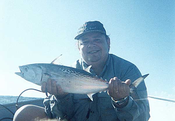 Man in a blue shirt and cap holds a striped fish, with a boat and ocean visible