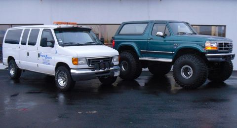 1996 Ford Bronco - Two vehicles, a modified Ford Bronco and a Ford van, are parked outside a building.