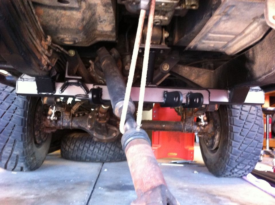 Undercarriage view of a vehicle's rear axle, driveshaft, and suspension components, with one tire visible