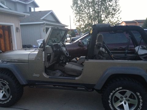 2005 Jeep Wrangler - A tan Jeep Wrangler is parked in front of a garage, with its doors off and a roll cage visible.
