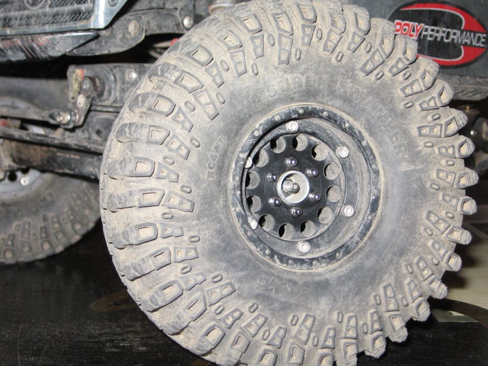 Close-up of a large, knobby off-road tire with a black beadlock wheel. A red and black 'POLY PERFORMANCE' sticker is visible in the background