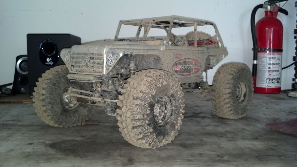Mud-covered off-road vehicle with large tires and roll cage, parked indoors next to a speaker and fire extinguisher