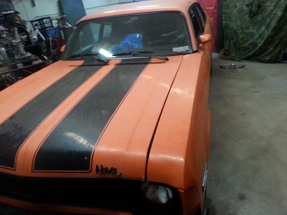 Orange 1973 Chevrolet Nova coupe with black racing stripes on hood and trunk, parked indoors
