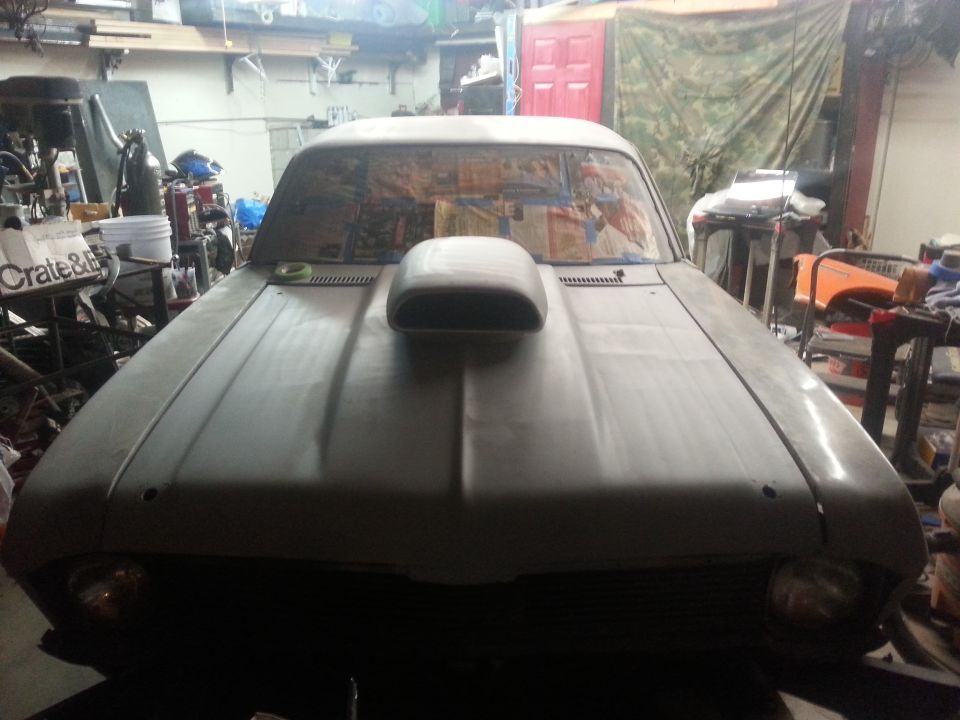 Car in garage, primer gray, with a large hood scoop. Windshield is covered with paper