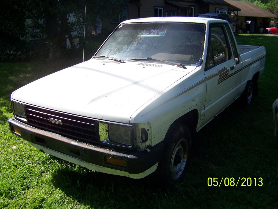 White Toyota pickup truck parked on grass, front passenger side visible. Has a 'For Sale' sign in the windshield