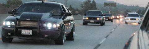 2003 Ford Crown Victoria - dark sedan fitted with a front push bar drives on a wet highway alongside other traffic at dusk.