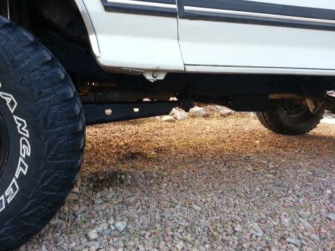 1991 Ford Bronco - The underside of a white truck, showing suspension components and a large tire, rests on a gravel surface.