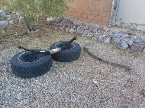 1991 Ford Bronco - Leaf springs and tires are laid out on a gravel surface with a brick wall and vegetation in the background.