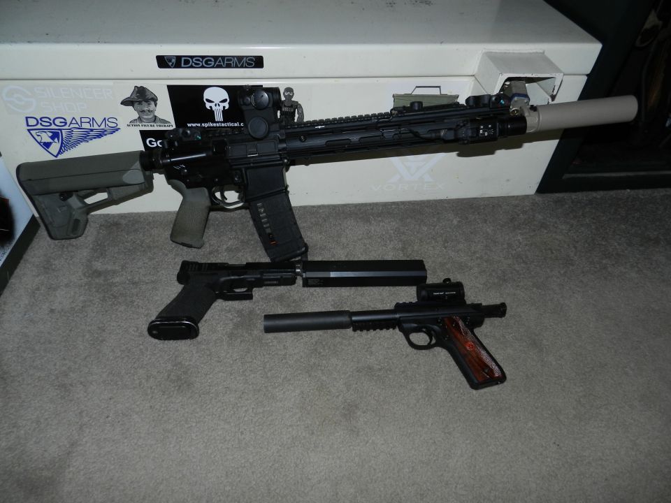 Assault rifle and two handguns with suppressors on a carpeted floor in front of a safe