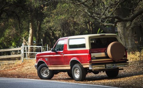 1994 Ford Bronco - A red and tan Ford Bronco is parked on the side of a road, surrounded by trees and foliage.