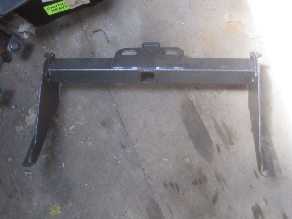 Trailer hitch receiver with mounting brackets on concrete floor. Sticker on black box reads 'CAUTION HEAVY'