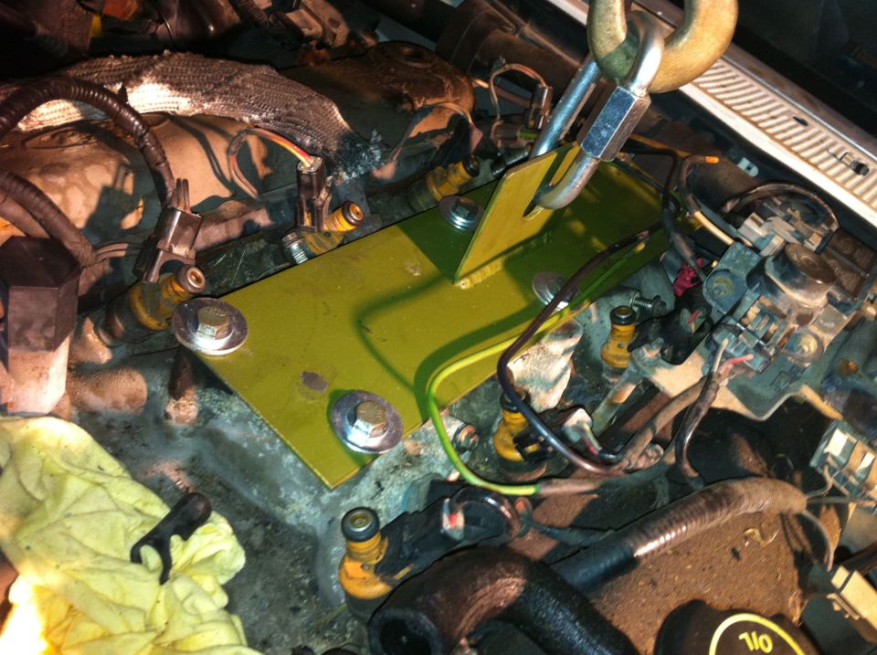 Engine bay with a yellow plate and bolts being installed, wires and hoses visible