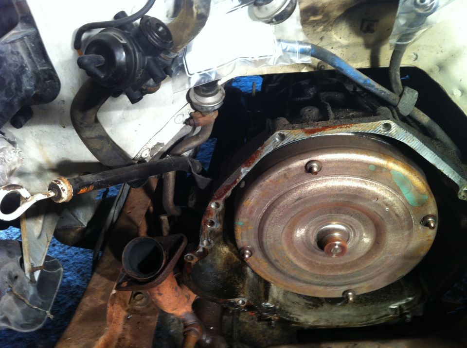 Close-up of a vehicle's transmission bell housing and torque converter, with various hoses and vacuum lines visible