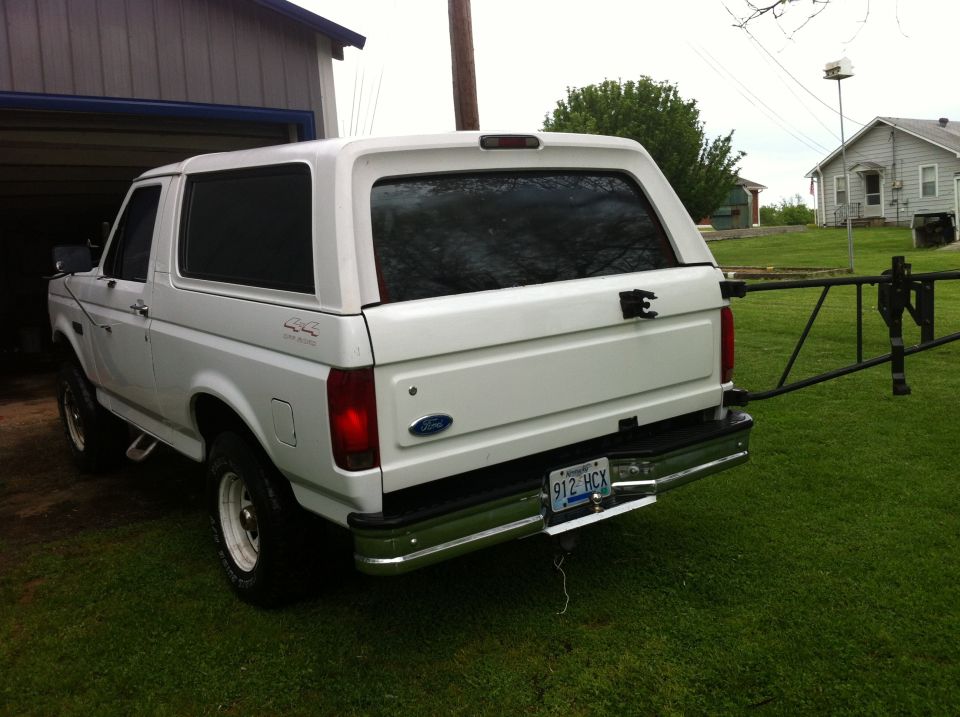 White 1992 Ford Bronco with a camper shell, rear view, parked on grass next to a garage