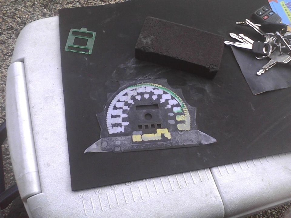 Car dashboard gauge cluster with white and green markings, keys, and a sanding block on a black surface