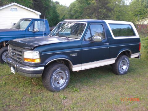 1993 Ford Bronco - A dark green and white Ford Bronco is parked on a grassy area next to a blue pickup truck.