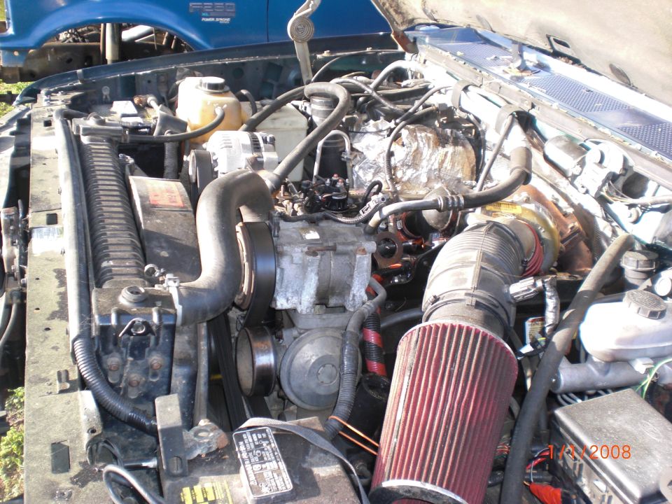 Engine bay with hoses, belts, and air filter visible. Blue truck in background