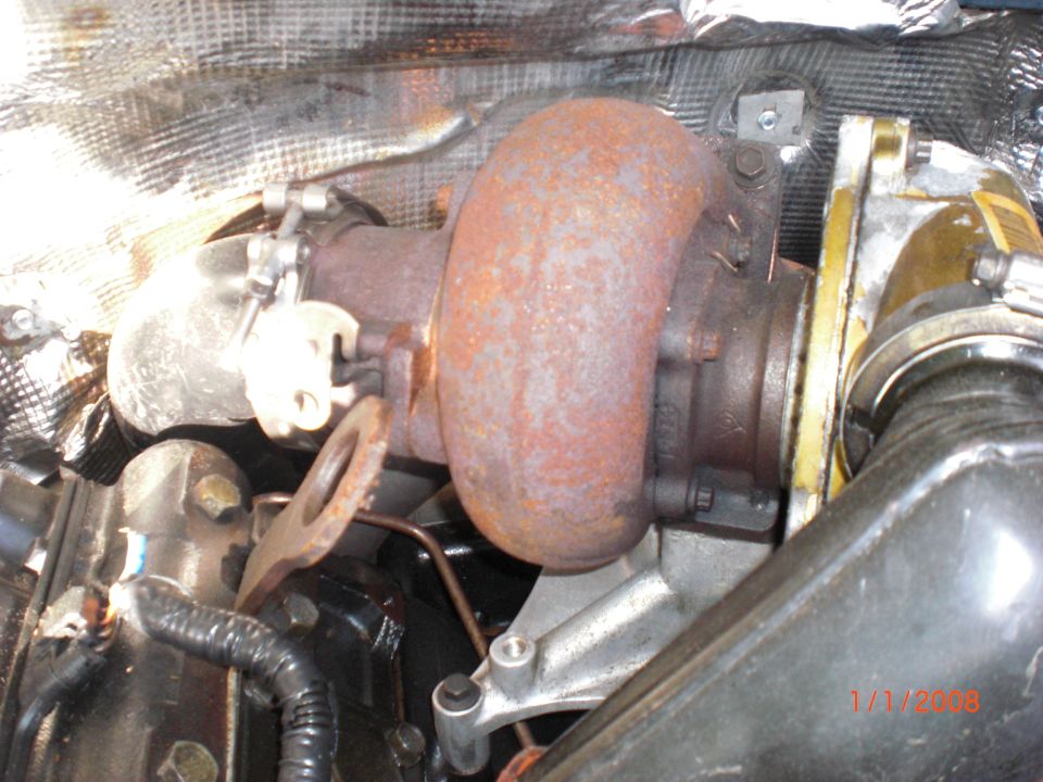Close-up of a rusty turbocharger with heat shielding above it. Some hoses and engine parts are visible below