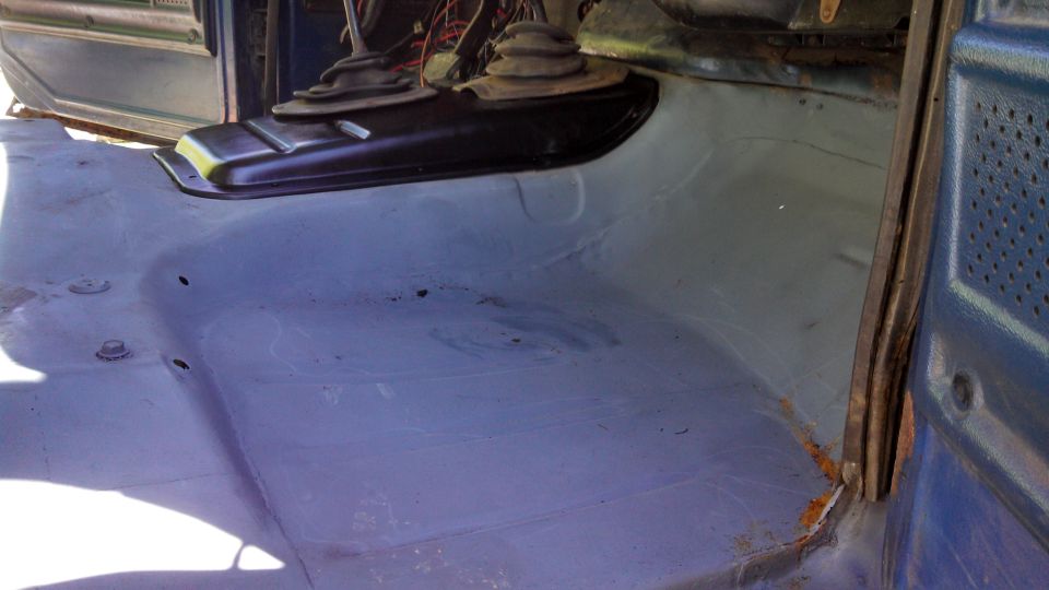 Interior floor of a vehicle, showing the transmission tunnel and bare metal floor. Some rust is visible near the door frame
