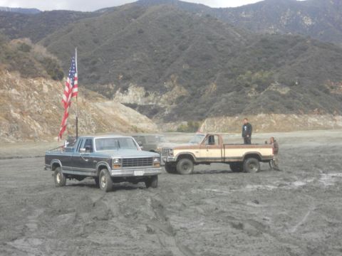 1986 Ford Bronco - Two pickup trucks are parked in a muddy area with hills in the background. An American flag flies from the blue truck.