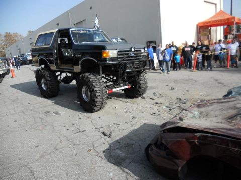 1986 Ford Bronco - A heavily modified black Ford Bronco with a tan top is parked next to a severely damaged car.