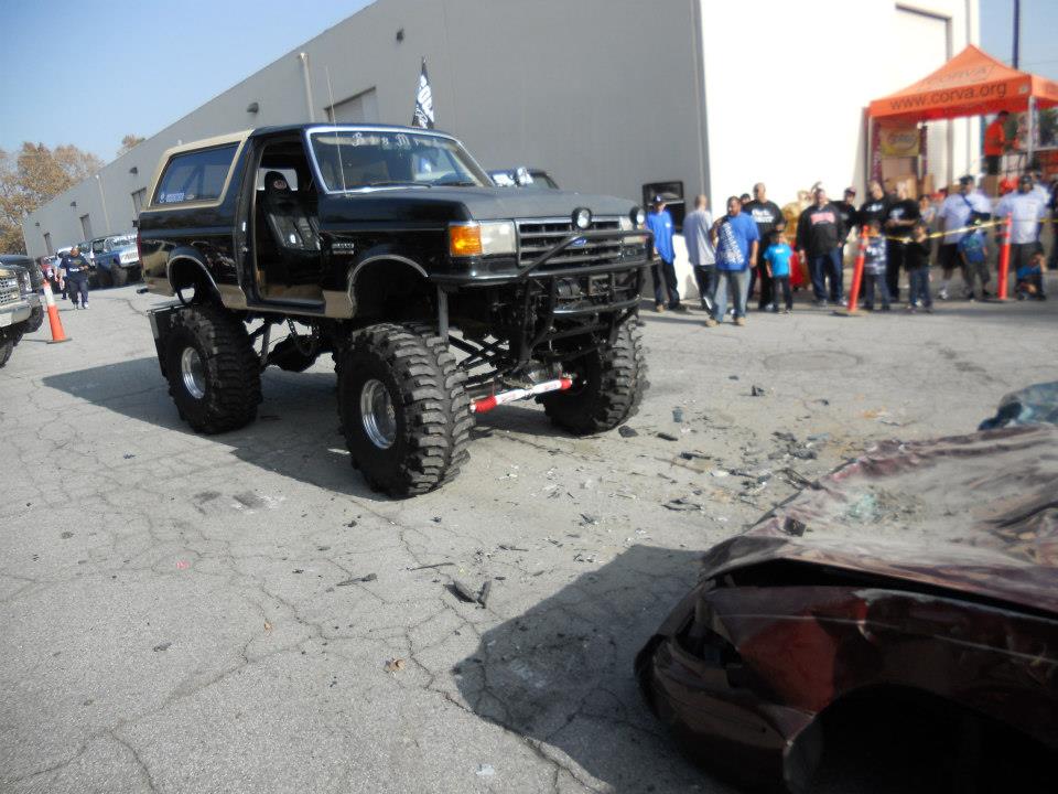 Lifted black Ford Bronco with a tan top, custom bumper, and large tires parked next to a crushed car