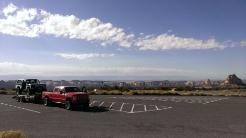 1994 Ford Bronco - A red pickup truck tows a trailer carrying a classic Ford Bronco through a scenic desert overlook.