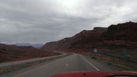1994 Ford Bronco - View from a red vehicle driving on a highway through a desert landscape with prominent red rock formations under a cloudy sky.