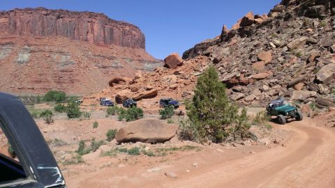 1994 Ford Bronco - Multiple off-road vehicles navigate a dusty trail amidst a desert canyon with towering red rock cliffs.