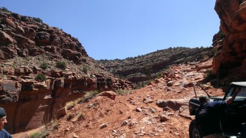 1994 Ford Bronco - Several off-road vehicles are seen traversing a challenging, rocky trail through a canyon with steep, red rock formations.