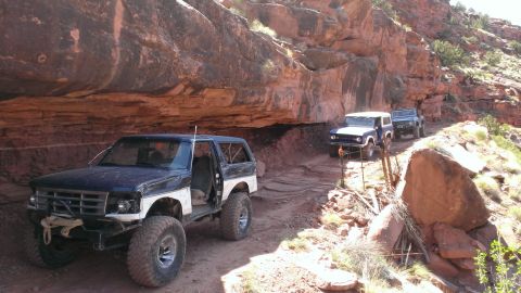 1994 Ford Bronco - Several SUVs, including a 1994 Ford Bronco, navigate a narrow dirt path through a canyon with towering rock formations.
