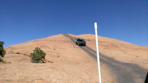 1994 Ford Bronco - A dark SUV ascends a dirt path on a large, arid rock formation under a bright blue sky.