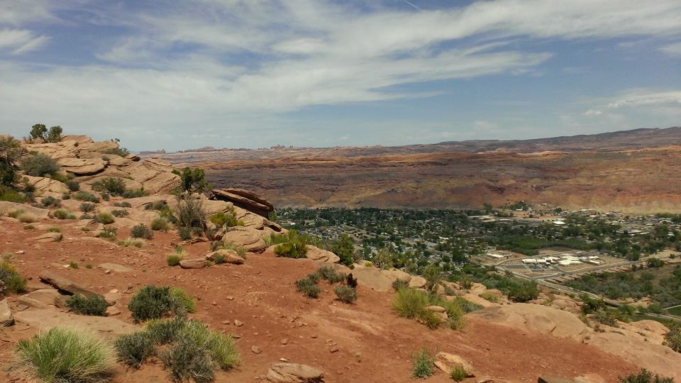Desert landscape overlooking a town, with red rocks and sparse vegetation in the foreground. Watermark reads www.supermotors.net