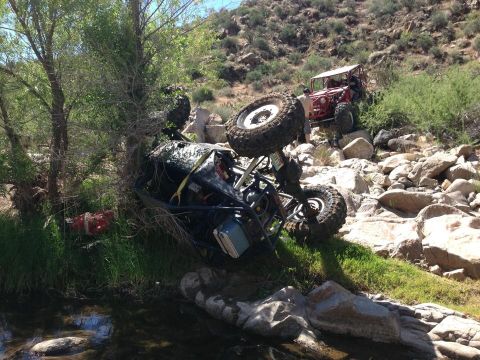1966 Ford Bronco - An off-road vehicle has overturned on a rocky incline near a stream, while another vehicle is visible further up the slope.