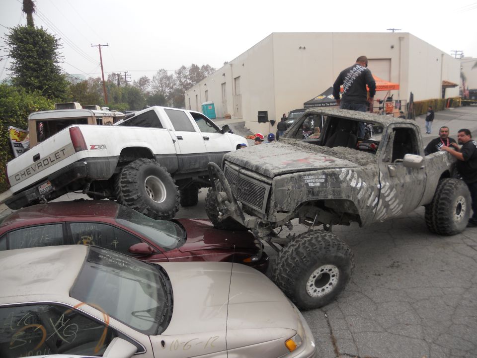 Mud-covered pickup truck with large tires, front grille visible, parked on other cars