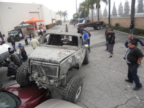 1986 Ford Bronco - A heavily modified truck, caked in mud, sits prominently in a parking lot with onlookers nearby spectators nearby.