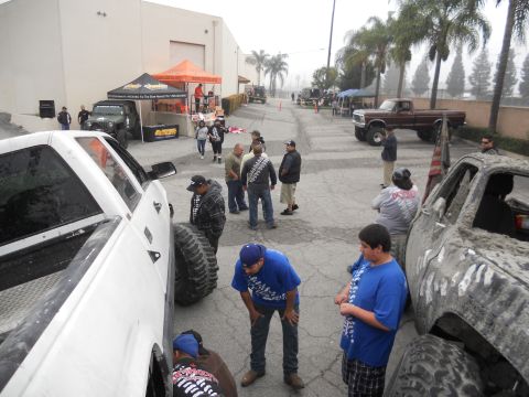 1986 Ford Bronco - People mill around modified trucks at an outdoor event, with tents and buildings in the background.