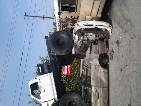 1986 Ford Bronco - A severely damaged white sedan is wedged under a large truck, with graffiti on the side of the truck.