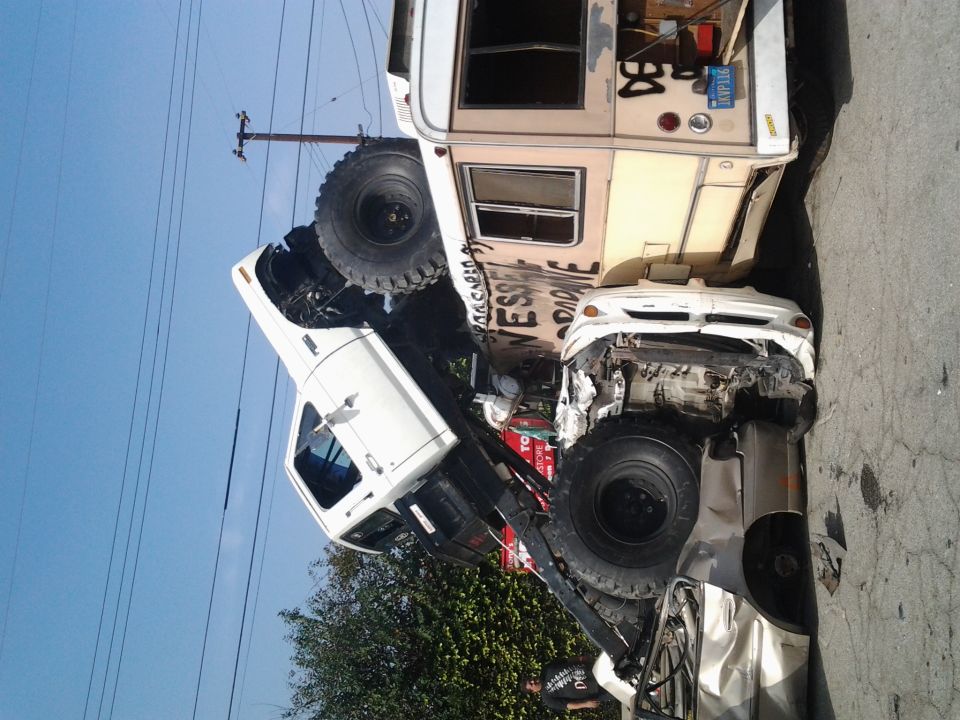 wrecked white truck on top of a silver car, with a camper van behind
