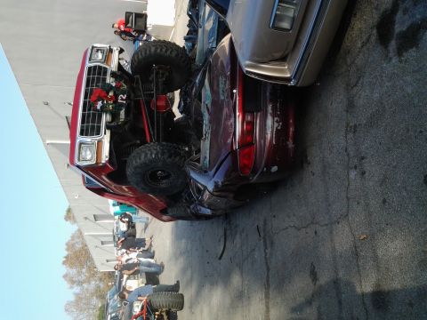 1986 Ford Bronco - A red lifted truck has rolled onto its side, crushing a red sedan beneath it.