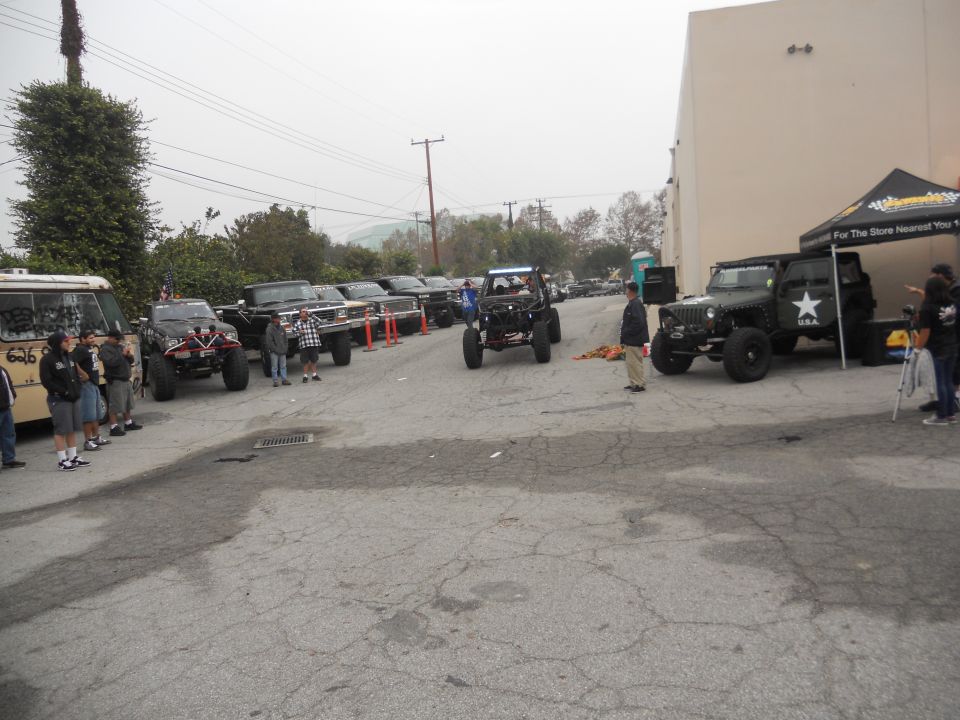 Several modified off-road vehicles and a van parked in a lot with people standing around