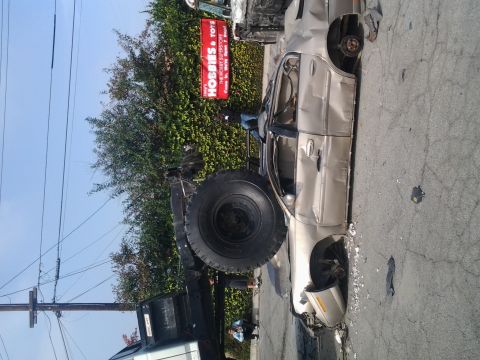 1986 Ford Bronco - A severely damaged gold sedan lies on its side, with a large truck and its oversized tire nearby.