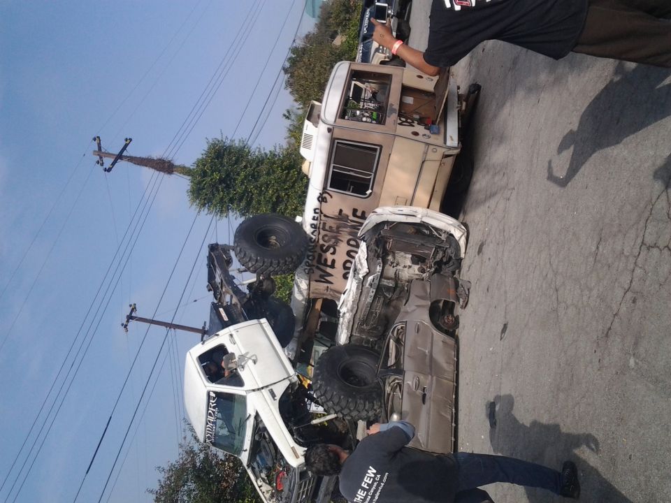 Two heavily modified off-road trucks are shown, one overturned on its side. Text on the side of the upright truck reads 'WESSEL GOODONE'