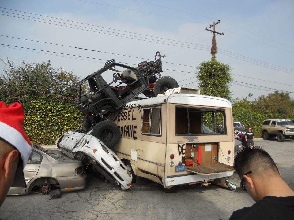 bus with buggy on top, wrecked cars underneath, people watching